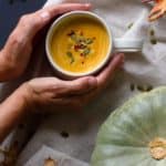 Two hands holding a white cup of pumpkin soup, cosy fall scene.