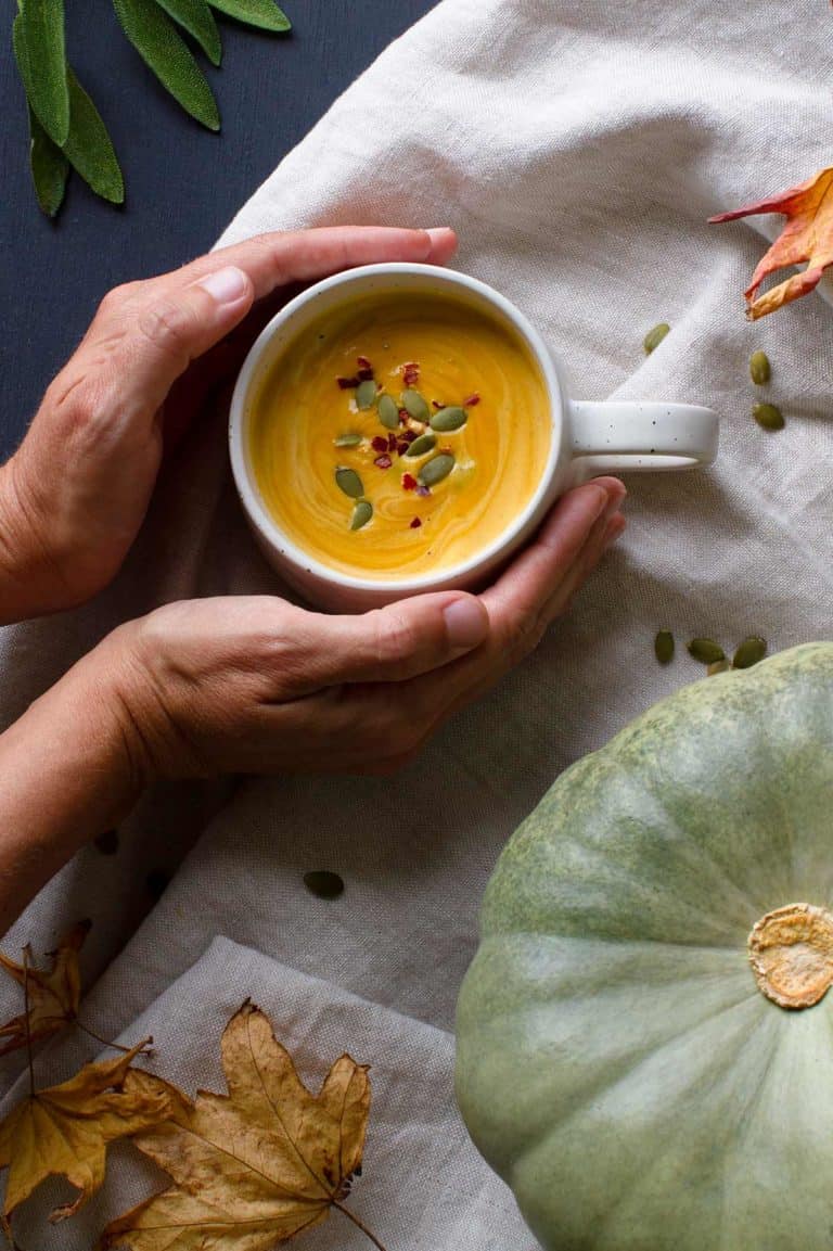 Two hands holding a white cup of pumpkin soup, cosy fall scene.