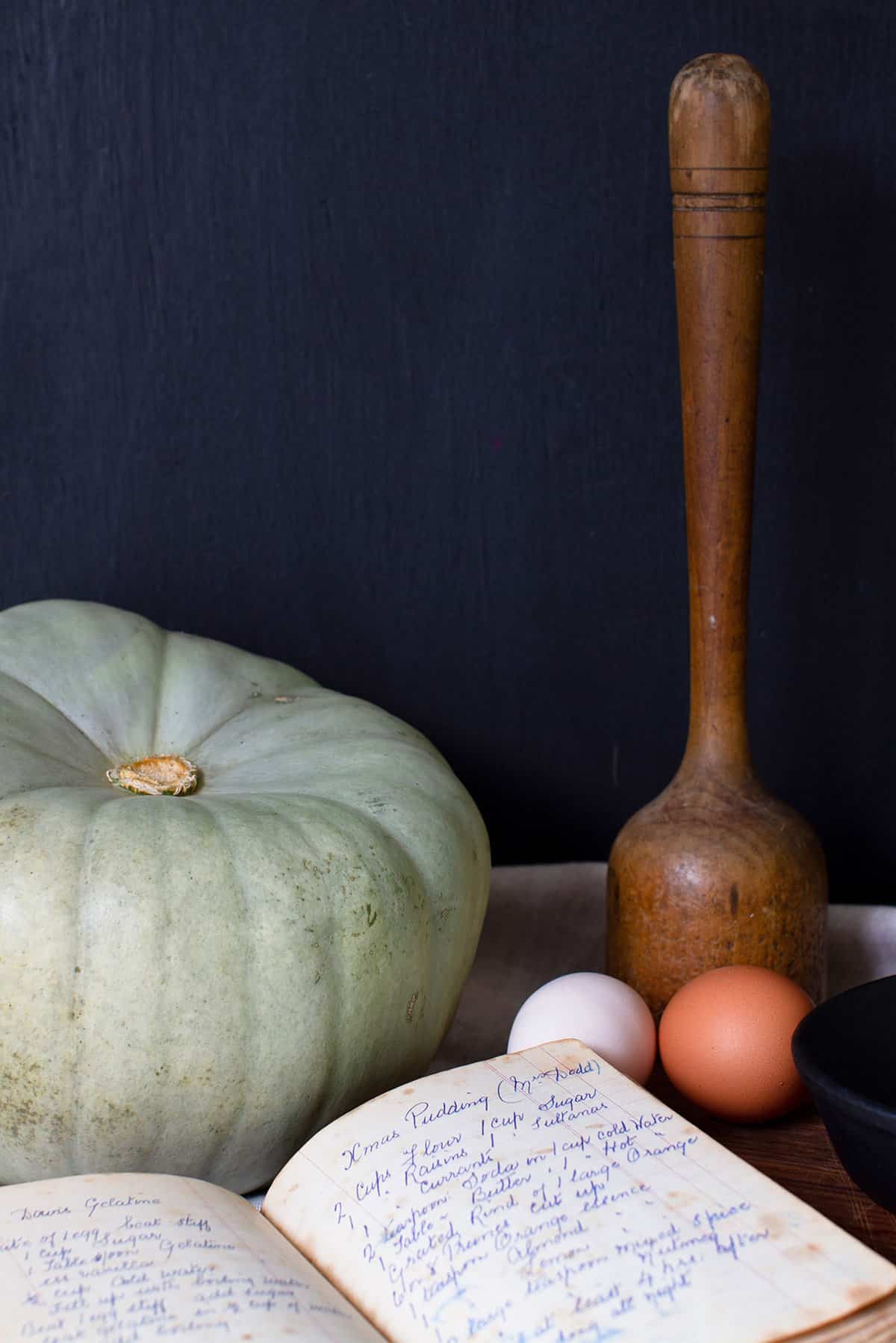 A moody kitchen vignette including a pumpkin, recipe book, eggs and a sauerkraut pounder