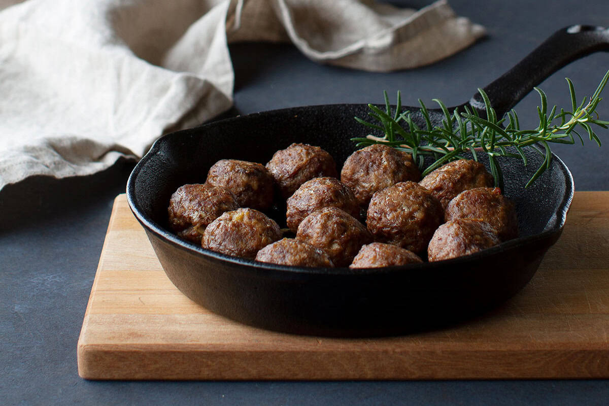 Cooked meatballs in a cast iron pan on a wooden board next to a linen tea towel.