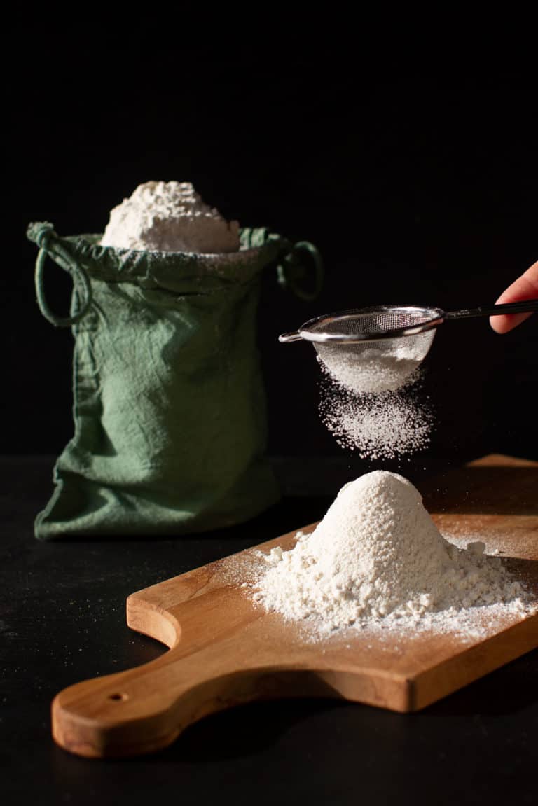 Flour shaped into a cone on a wooden board. Green linen bag with flour in the background. Moody scene.