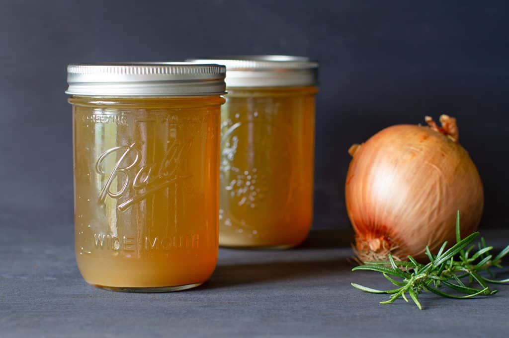 Two jars of golden lamb stock with an onion and sprig of rosemary. Moody scene