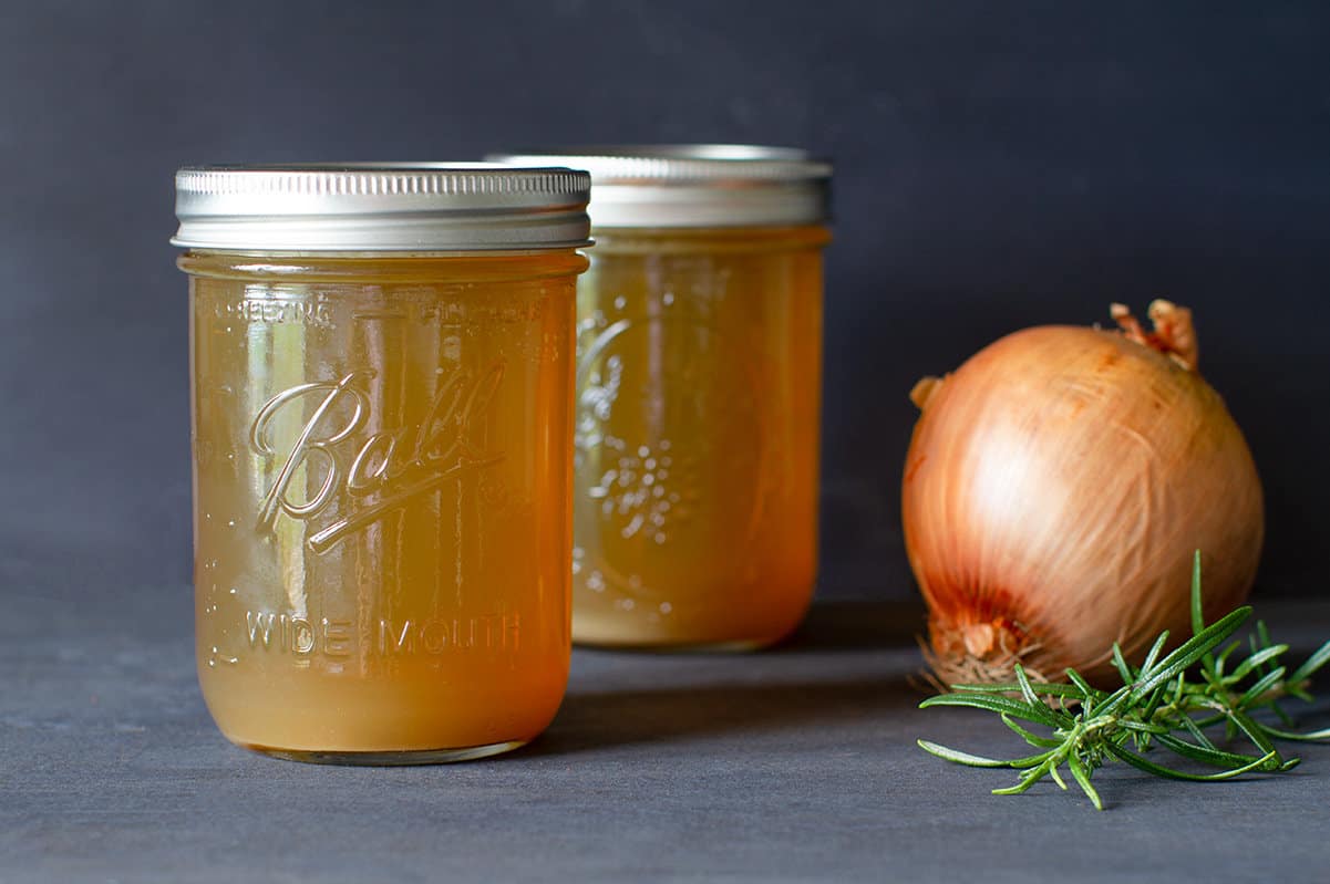 Two jars of golden lamb stock with an onion and sprig of rosemary. Moody scene