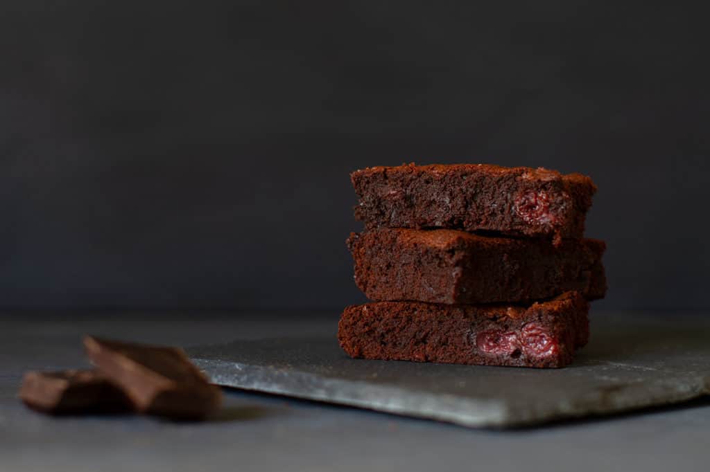 Three stacked squares of cherry brownies on a shingle next to a square of dark chocolate. Dark background.