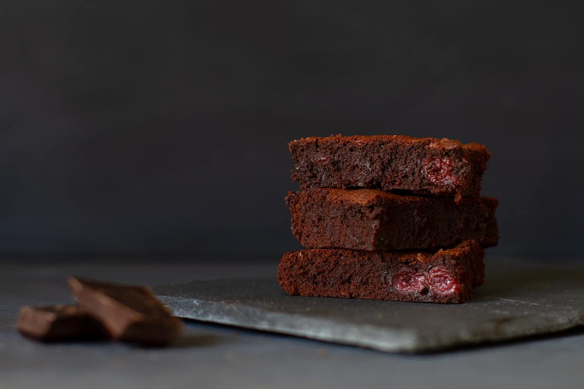 Three stacked squares of American brownies on a shingle next to a square of dark chocolate. Dark background.