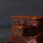 Three stacked squares of American brownies on a shingle next to a square of dark chocolate. Dark background.