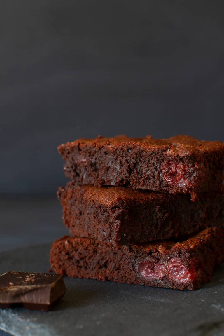 Three stacked squares of American brownies on a shingle next to a square of dark chocolate. Dark background.