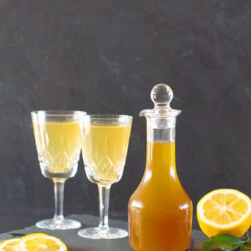 A glass jar of lemon cordial next to two vintage wine glasses with lemonade on a black shingle tile.