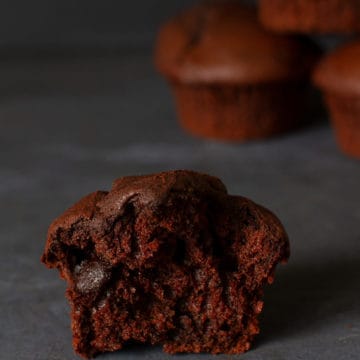 Close-up of a half triple chocolate muffin next to some chocolate drops and a stack of blurred muffins in the back.