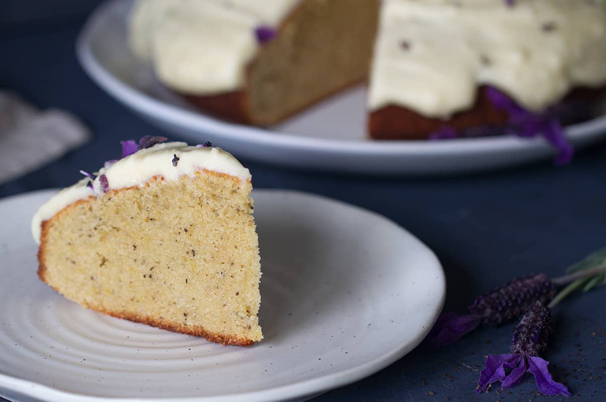 A slice of earl grey lavender cake on a white plate next to lavender. The full cake is blurred out in the background.