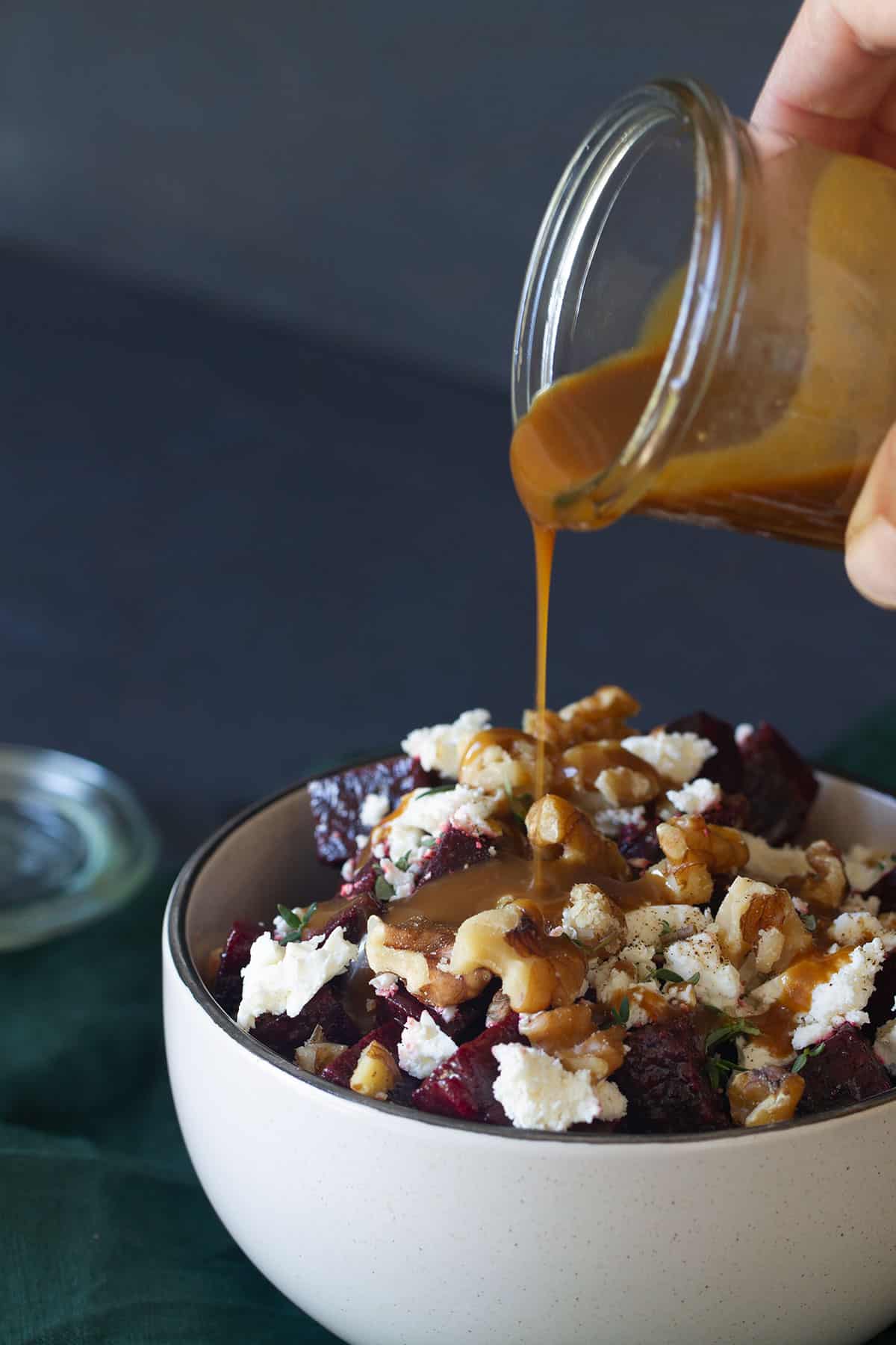 A bowl of roasted beetroot walnut and feta salad on a green tea towel. A jar of dressing is being poured onto the salad.