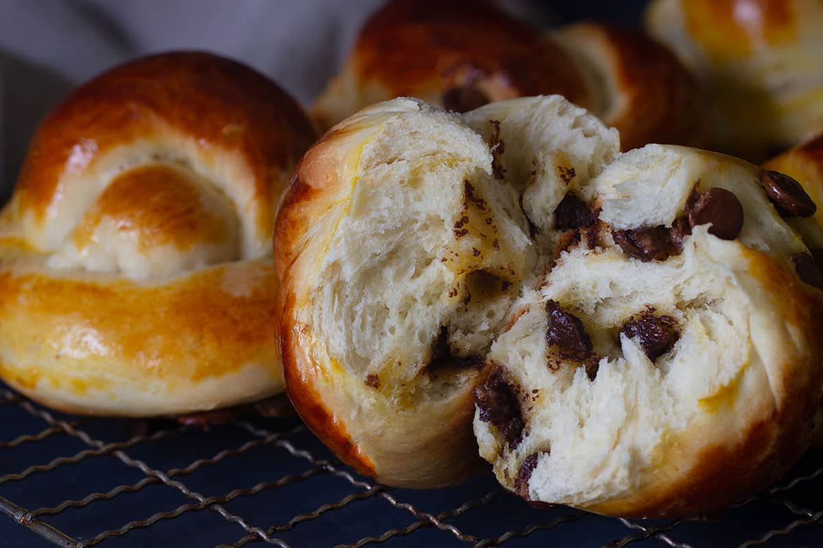 Close-up of a chocolate knotted bun on a cookie rack. A couple of buns and a linen tea towel are blurred out in the background.