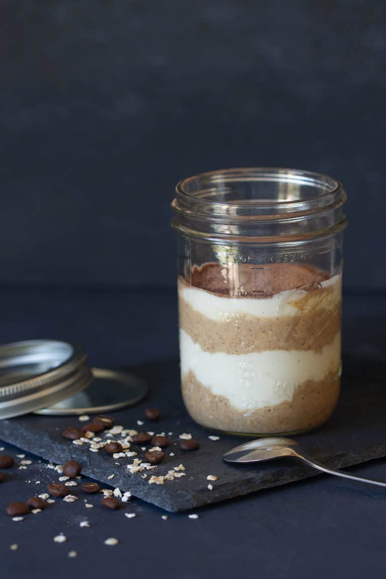 A clear jar of tiramisu overnight oats on a shingle plate. Mason jar lid, spoon, oats, and coffee beans are next to it.