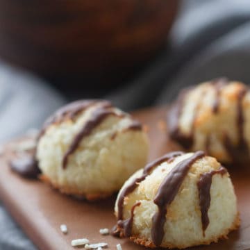 Three coconut macaroons (Kokosmakronen) on a wooden board and grey tea towel. A bowl of extra macaroons is blurred out in the background.