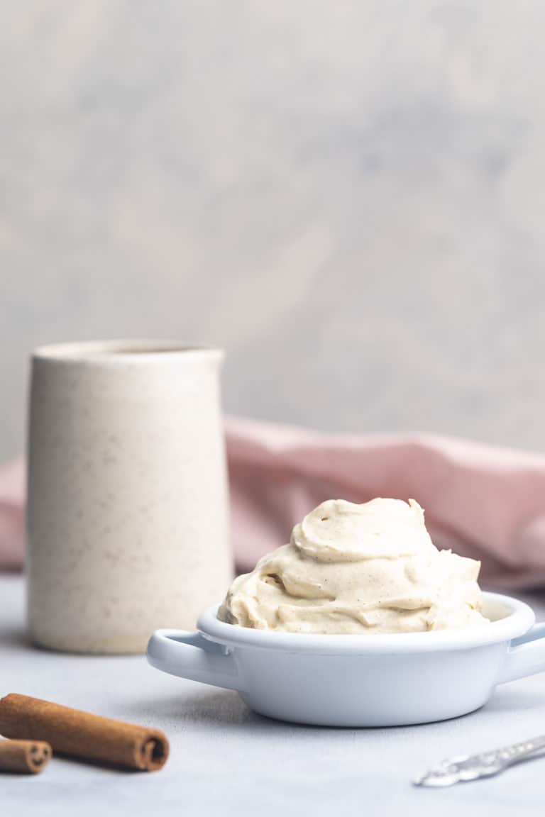 A bowl of whipped chantilly mascarpone next to a white ceramic mug, pink napkin and cinnamon sticks.