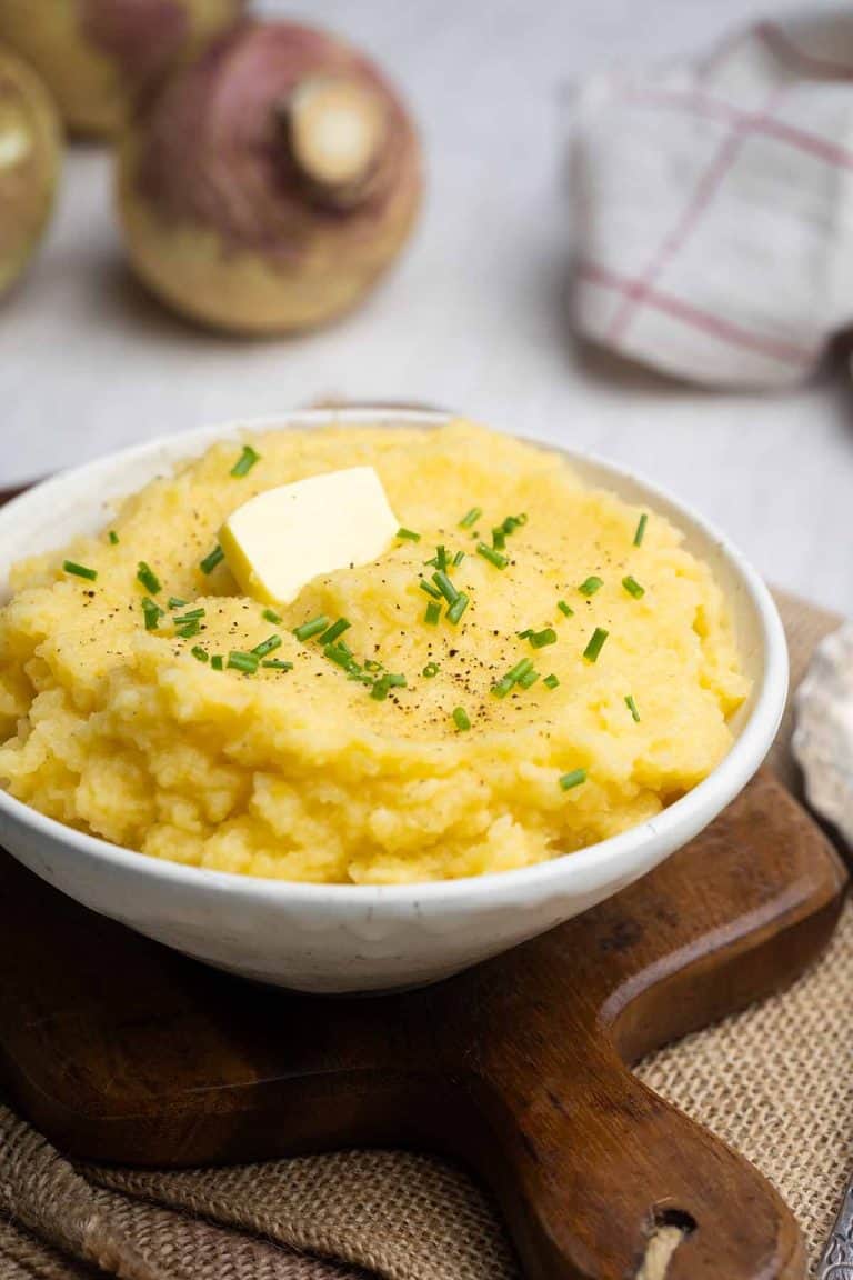 A bowl of extra creamy swede mash on a wooden board, next to swedes and a tea towel.