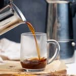 A Moka pot pouring coffee into a glass cup.