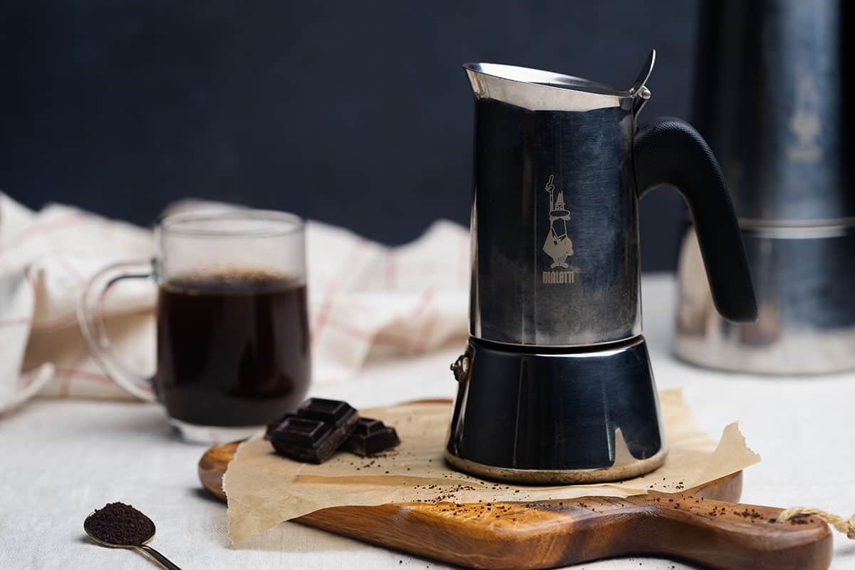 A Moka pot on a wooden board next to a cup and another larger Moka Pot.