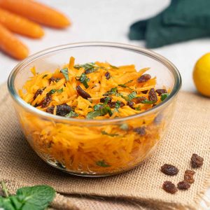 A glass bowl filled with raw carrot salad, garnished with mint and raisins.