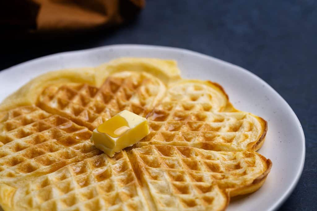 A fluffy American waffles on a plate being drizzled with maple syrup.