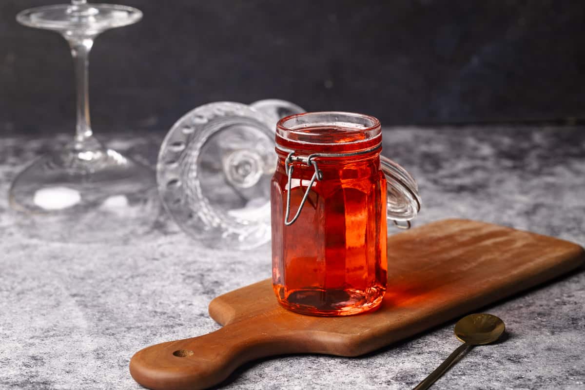 A jar of homemade Rosé simple syrup on a wooden board next to wine glasses.