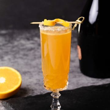A glass of Rosé mimosa on a slate next to an orange and a bottle of Rosé champagne.