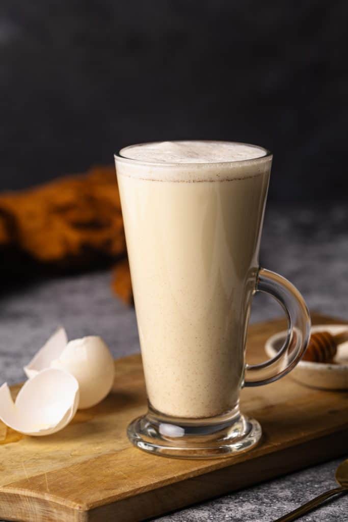 A tall glass cup filled with creamy egg milk on a wooden board next to egg shells.