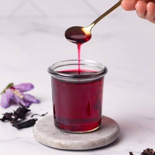 Homemade hibiscus syrup in a glass jar. A spoon is scooping some syrup.