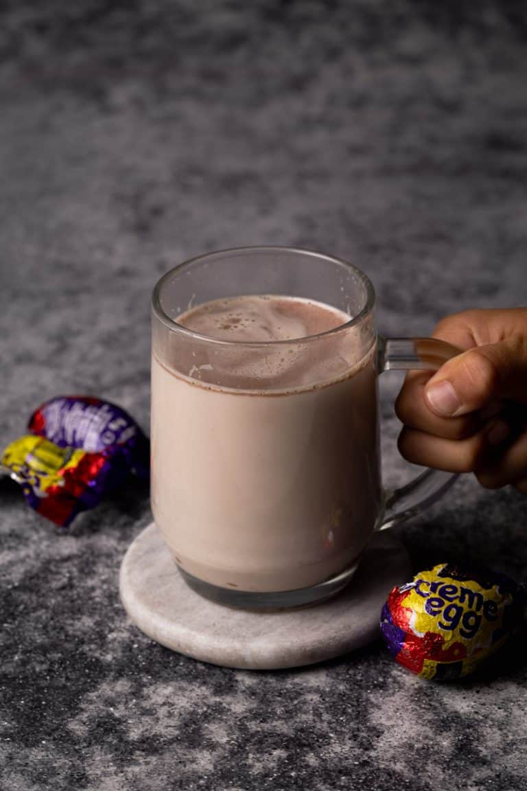 Cadbury creme egg hot chocolate in a glass cup next to creme eggs.