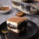 A slice of brownie tiramisu on a black plate next to the whole dish.