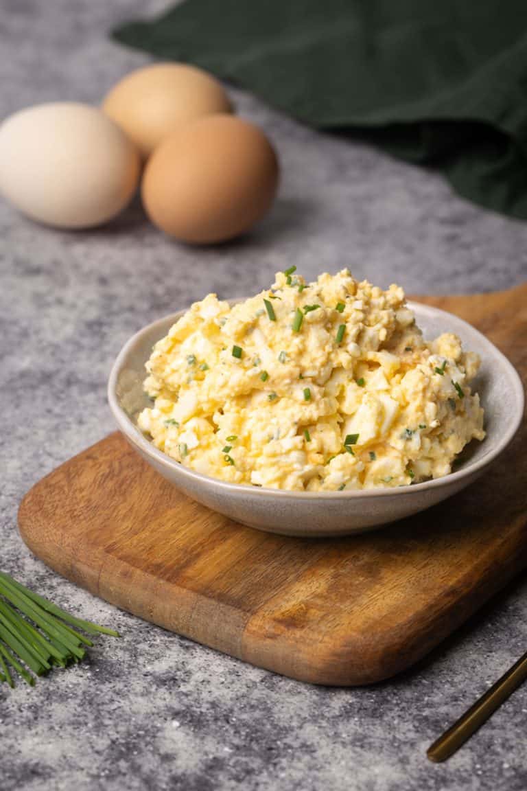 Egg salad in a bowl on a wooden board next to eggs and a green napkin.