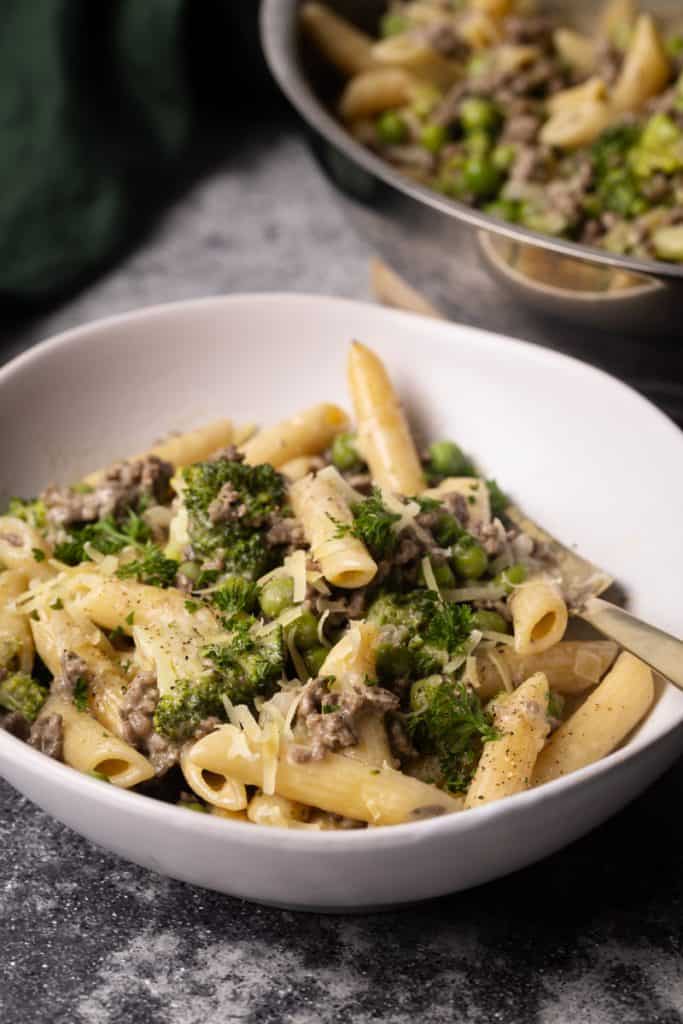 A white bowl filled with ground beef Alfredo pasta next to a large pan.
