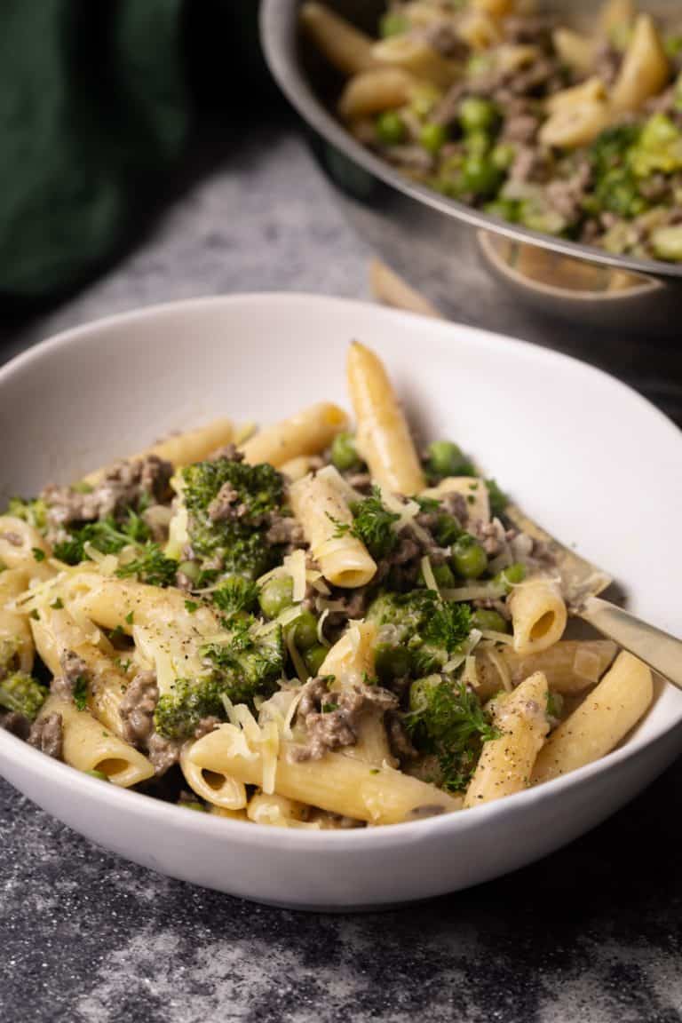 A white bowl filled with ground beef Alfredo pasta next to a large pan.