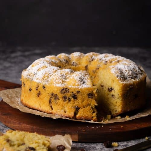 A raisin cake on a wooden board next to a piece of cake on some parchment paper.