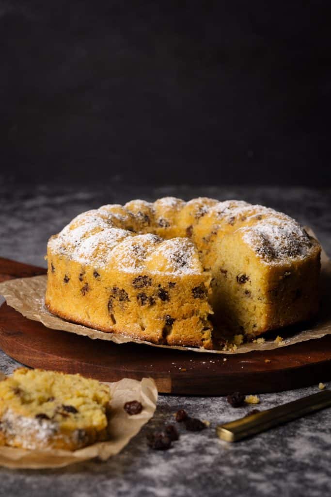 A raisin cake on a wooden board next to a piece of cake on some parchment paper.