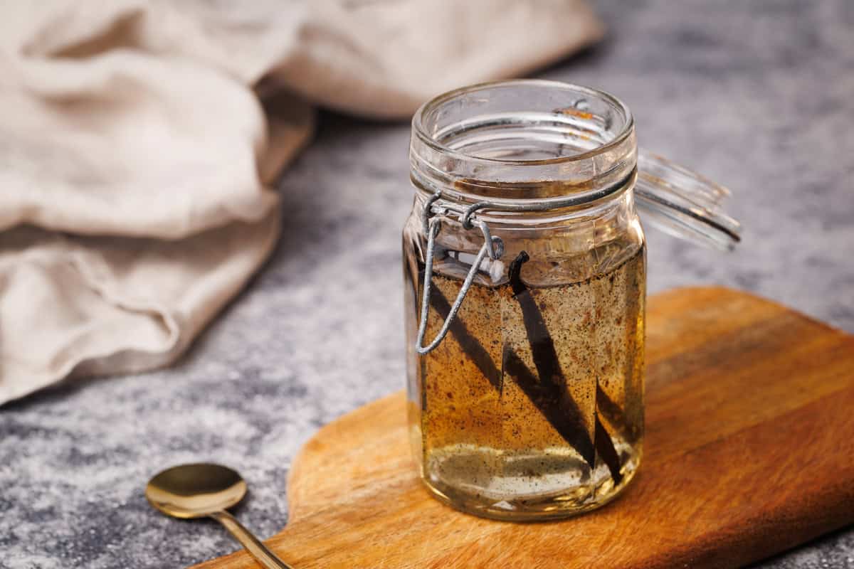 Vanilla syrup in a glass container on a wooden board next to a golden spoon and a linen napkin.