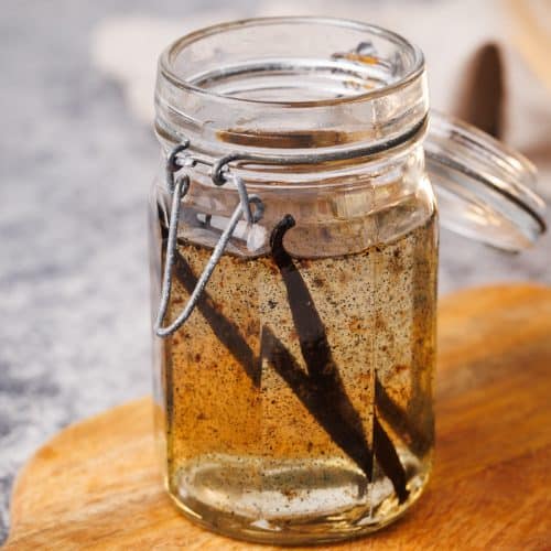 Vanilla syrup in a glass container on a wooden board next to a golden spoon and a linen napkin.