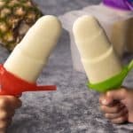 Tropical popsicles held by kids' hands next to a pineapple and ice block moulds.