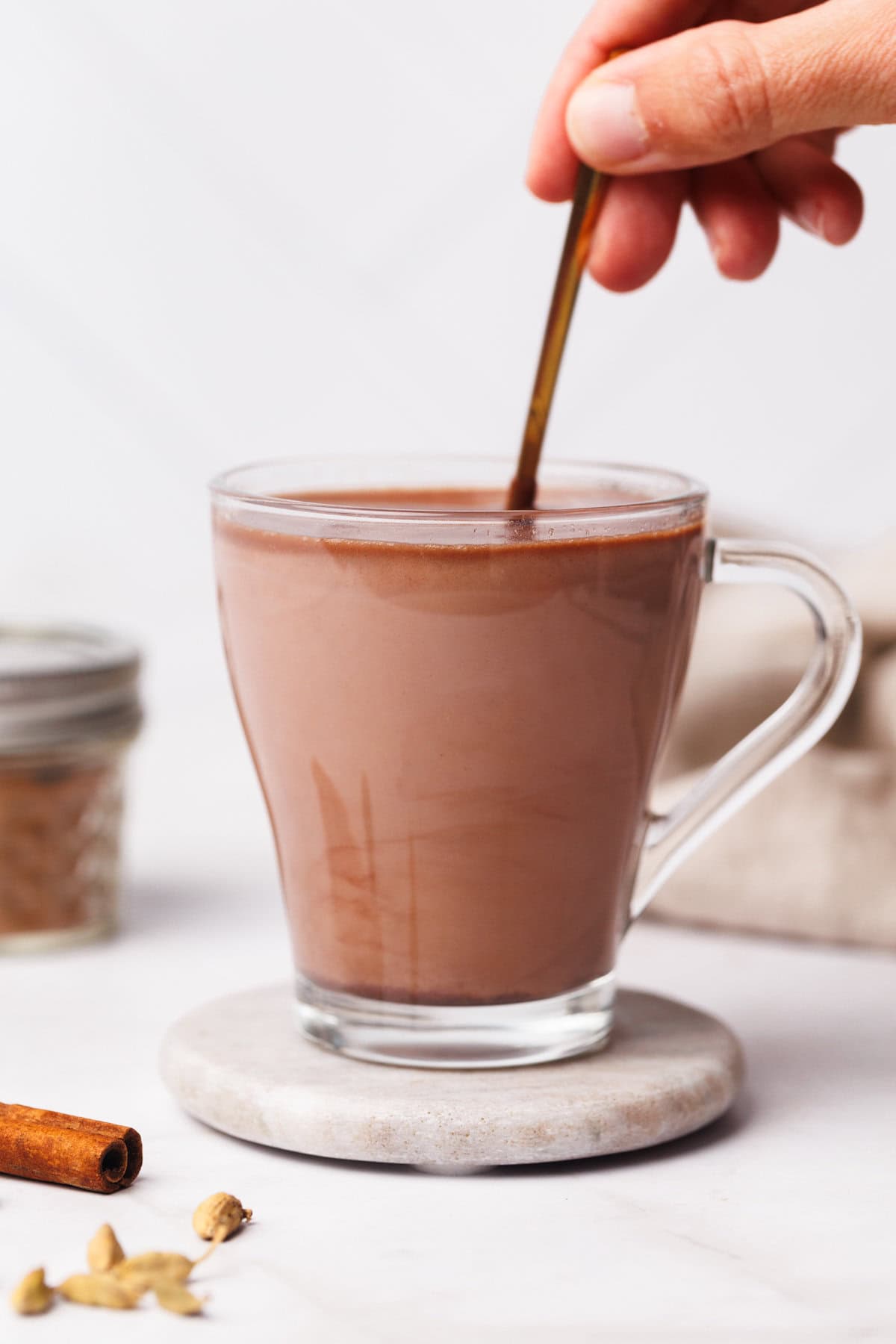 A glass cup filled with chai hot chocolate on a marble coaster next to a jar of spice mix and a linen tea towel.