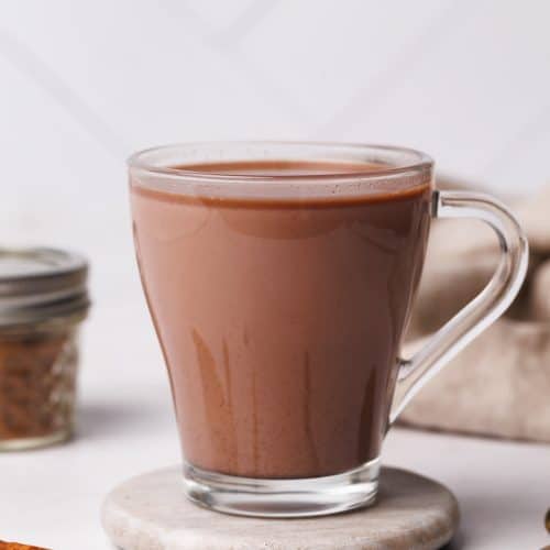 A glass cup filled with chai hot chocolate on a marble coaster next to a jar of spice mix and a linen tea towel.