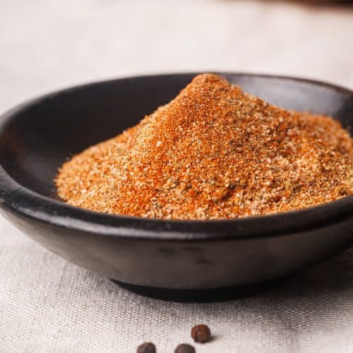 Lamb dry rub in a black bowl next to pepper kernels.