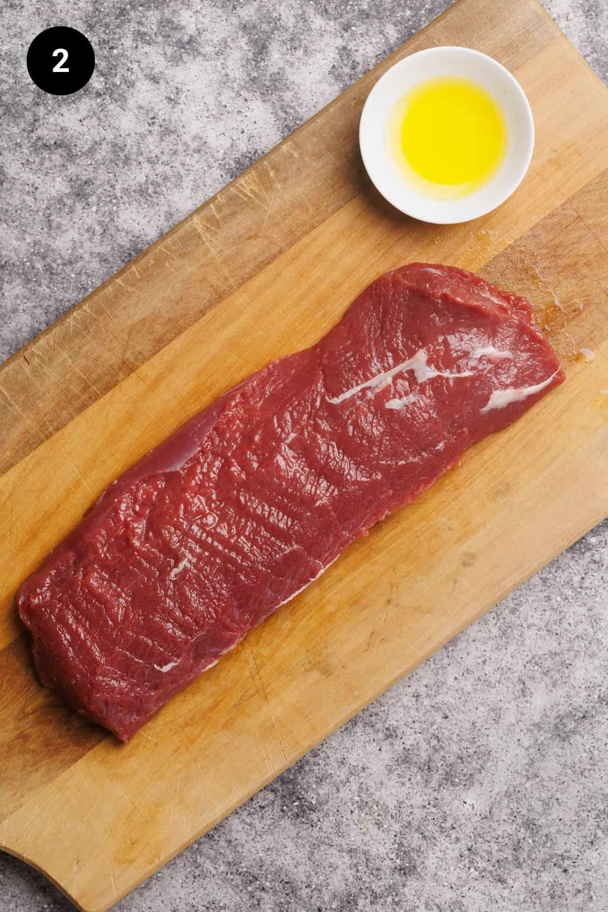 Lamb loin and a small bowl of olive oil on a wooden board.