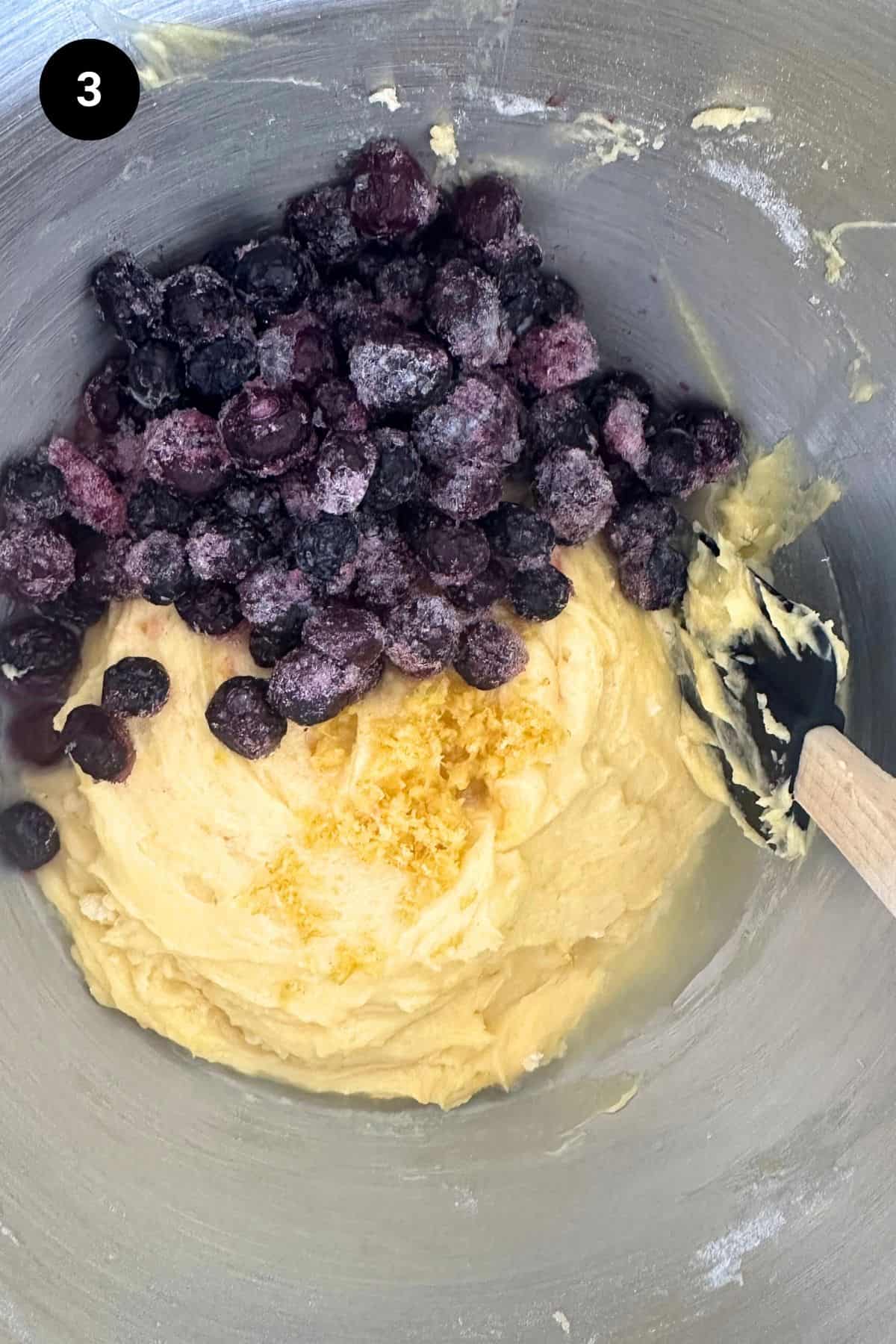 Blueberries and lemon zest on top of the cake batter in a metal bowl.