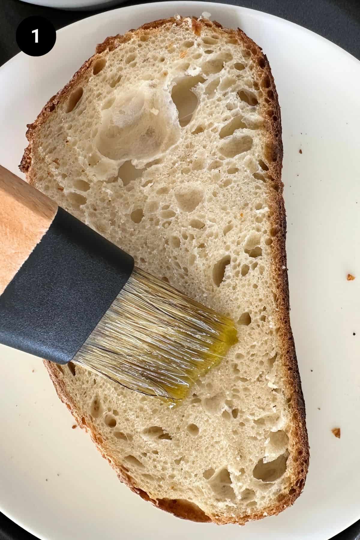 Bread slice on a white plate being brushed with olive oil.