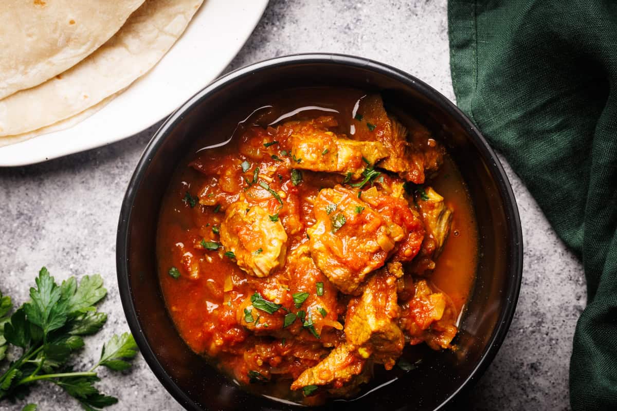A black bowl filled with lamb curry next to naan bread and a green napkin.