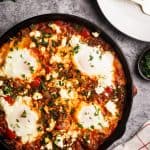 Sausage shakshuka in a cast iron pan next to a tea towel, plates, and parsley.
