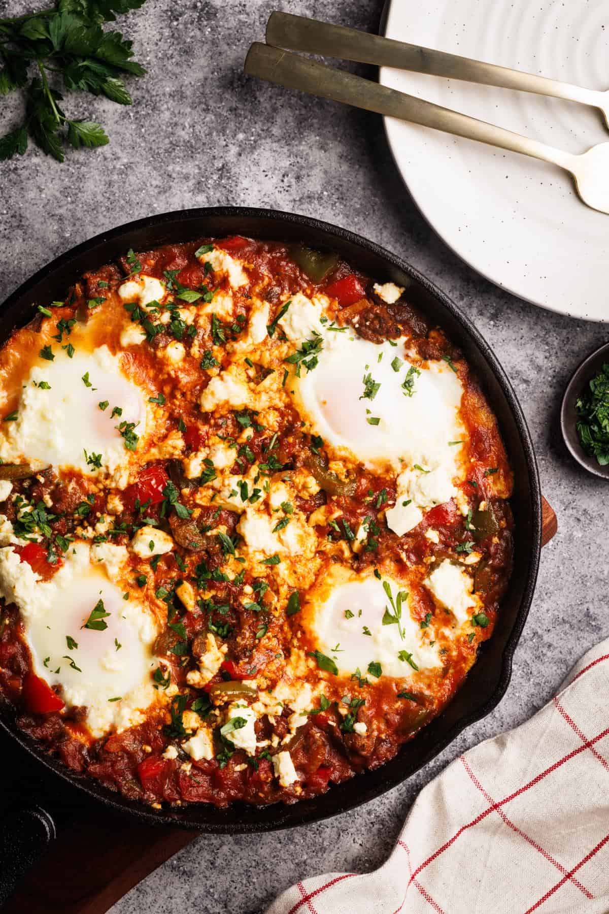Lamb shakshuka in a cast iron pan next to a tea towel, plates, and parsley.