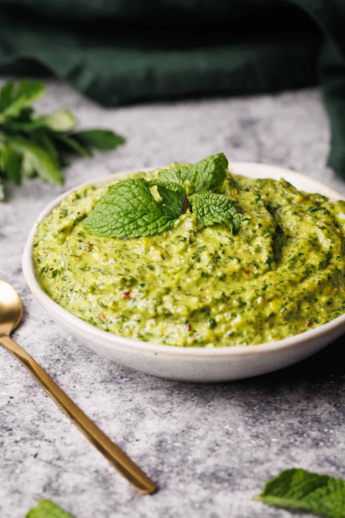 Mint pesto in a small white bowl next to a green napkin and golden spoon.