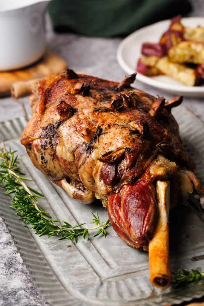 A roasted leg of lamb on a metal plate next to gravy and fresh rosemary.