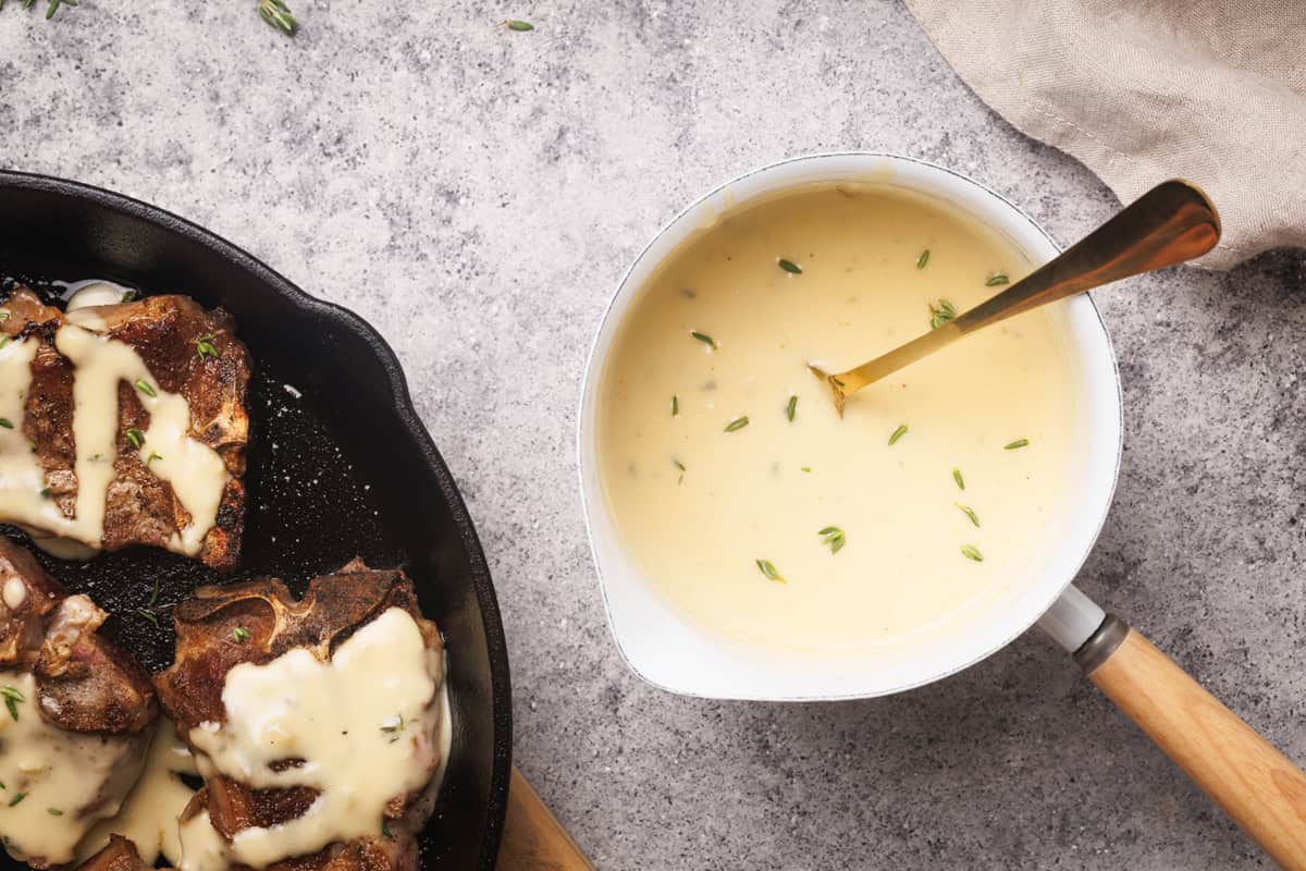 A saucepan with a creamy sauce next to lamb loin chops in a cast iron skillet.